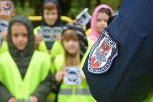 Zdjęcie kolorowe. Zbliżenie naszywki na ramieniu policyjnego munduru w tle dzieci z ulotkami mistrzu zwolnij