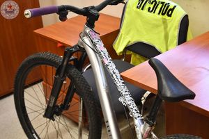 Zdjęcie kolorowe. Rower koloru szarek w tle przewieszona przez krzesło kamizelka odblaskowa z napisem policja