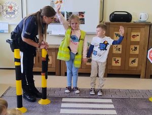 Zdjęcie kolorowe. Policjantka przeprowadza przedszkolaków przez matę na której jest namalowane przejście dla pieszych