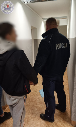 Zdjęcie kolorowe. Policjant doprowadza zatrzymanego mężczyznę