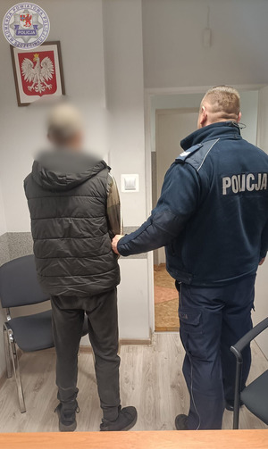 Zdjęcie kolorowe. Policjant doprowadza zatrzymanego mężczyznę