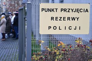 Zdjęcie kolorowe. Na ogrodzeniu przy bramie wejściowej tabliczka z napisem punkt przyjęcia rezerwy policji
