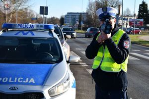 Zdjęcie kolorowe. Policjant przy radiowozie mierzy prędkość przejeżdżających pojazdów