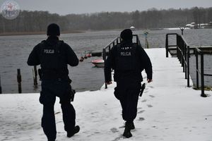 Zdjęcie kolorowe. Policjanci patrolują zaśnieżony teren przyległy do jeziora.