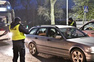 Zdjęcie kolorowe. Na drodze stoi dwóch policjantów. Jeden z nich zatrzymuje samochód do kontroli. Z lewej strony wjeżdża na jezdnię autobus.