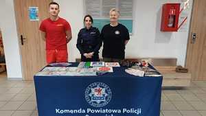 Zdjęcie kolorowe. Za stołem z logo szczecineckiej policji stoi od lewej ratownik wodny, policjantka i przedstawiciel sanepidu