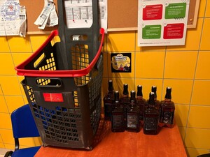 Kolorowe zdjęcie. Na zdjęciu w centralnej części stoi plastikowy wózek sklepowy obok którego z prawej strony stoi 8 butelek z płynem ciemnego koloru. w tle tablica informacyjna z poprzyczepianymi kartkami i broszurami