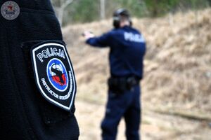 Zdjęcie kolorowe. Z lewej strony widoczna naszywka instruktora strzelań umieszczona na rękawie bluzy w tle strzelający policjant