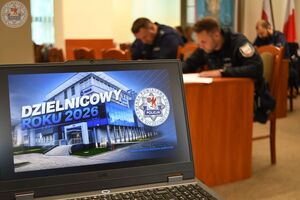 Zdjęcie kolorowe. Na pierwszym planie laptop z wyświetlonym zdjęciem budynku komendy w tle piszący test dzielnicowi
