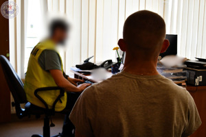 Zdjęcie. Na krześle siedzi odwrócony plecami mężczyzna przed nim przy biurku siedzi policjant w kamizelce odblaskowej