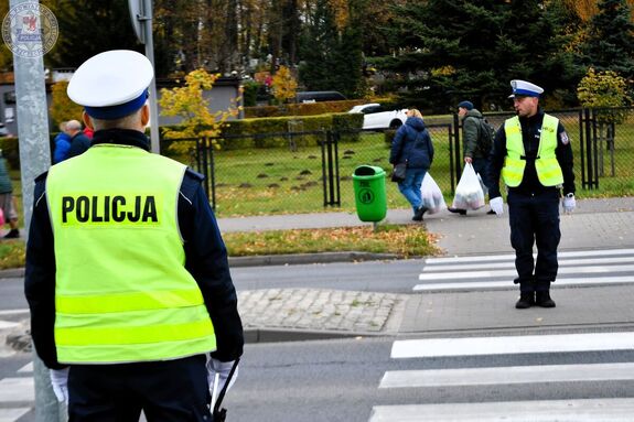 Zdjęcie kolorowe. Policjanci ruchu drogowego w pobliżu przejścia dla pieszych. Jeden z nich stojący na wysepce pomiędzy przejściami kieruje ruchem