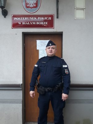 Zdjęcie kolorowe. Starszy sierżant Przemysław Romańczuk na tle drzwi wejściowych do Posterunku Policji w Białym Borze. W tle widać czerwoną tabliczkę z nazwą posterunku.