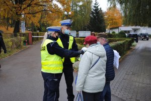 Zdjęcie kolorowe. Na zdjęciu widać jak policjanci z drogówki wręczają osobom wychodzącym lub wchodzącym na cmentarz opaski odblaskowe.