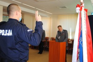 Zdjęcie kolorowe. Na pierwszym planie widać policjanta z uniesioną prawą dłonią do ślubowania. Z prawej strony widać sztandar Komendy Powiatowej Policji w Szczecinku. W oddali widać komendanta szczecineckich funkcjonariuszy, który odczytuje słowa roty.