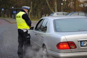 Zdjęcie kolorowe. Policjant rozmawia z kierującym, którego zatrzymał do kontroli drogowej. Kierujący siedzi w samochodzie koloru srebrnego.