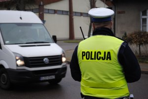 Zdjęcie kolorowe. Z lewej strony w kadrze umieszczony biały bus z prawej strony policjant obserwujący zachowania kierujących na drodze