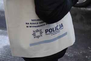 Bawełniana torba przewieszona przez ramię koloru ecri z niebieskim napisem Szczecinecka Fundacja na Rzecz Osób Poszkodowanych w Wypadkach Komunikacyjnych oraz Policja Komenda Powiatowa Policji w Szczecinku. Pod napisami znajduje się odblaskowy długi pasek.